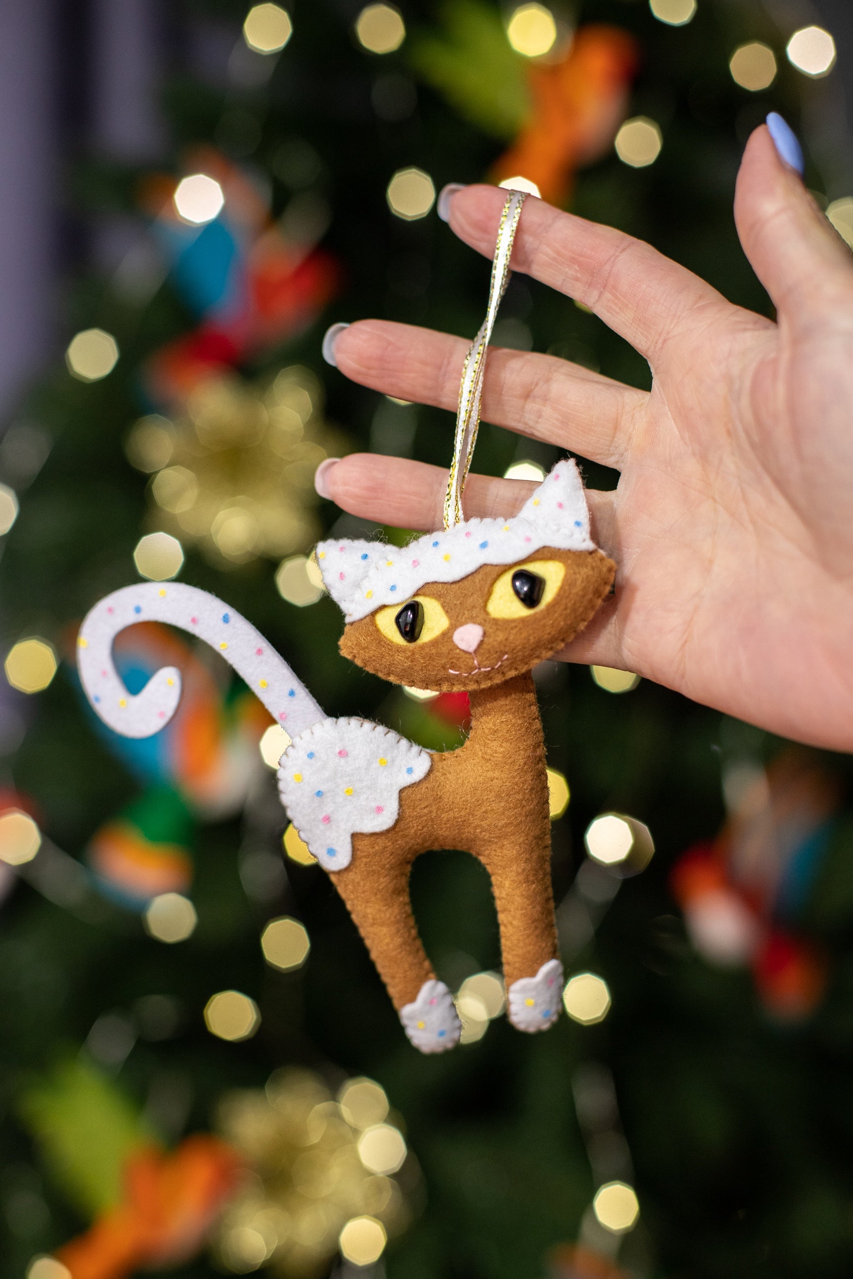 A handcrafted brown felt cat with white paws, chest, and a white tail. The cat has large yellow eyes and white felt pieces with multi-colored polka dots resembling icing on its head, paws and tail.
