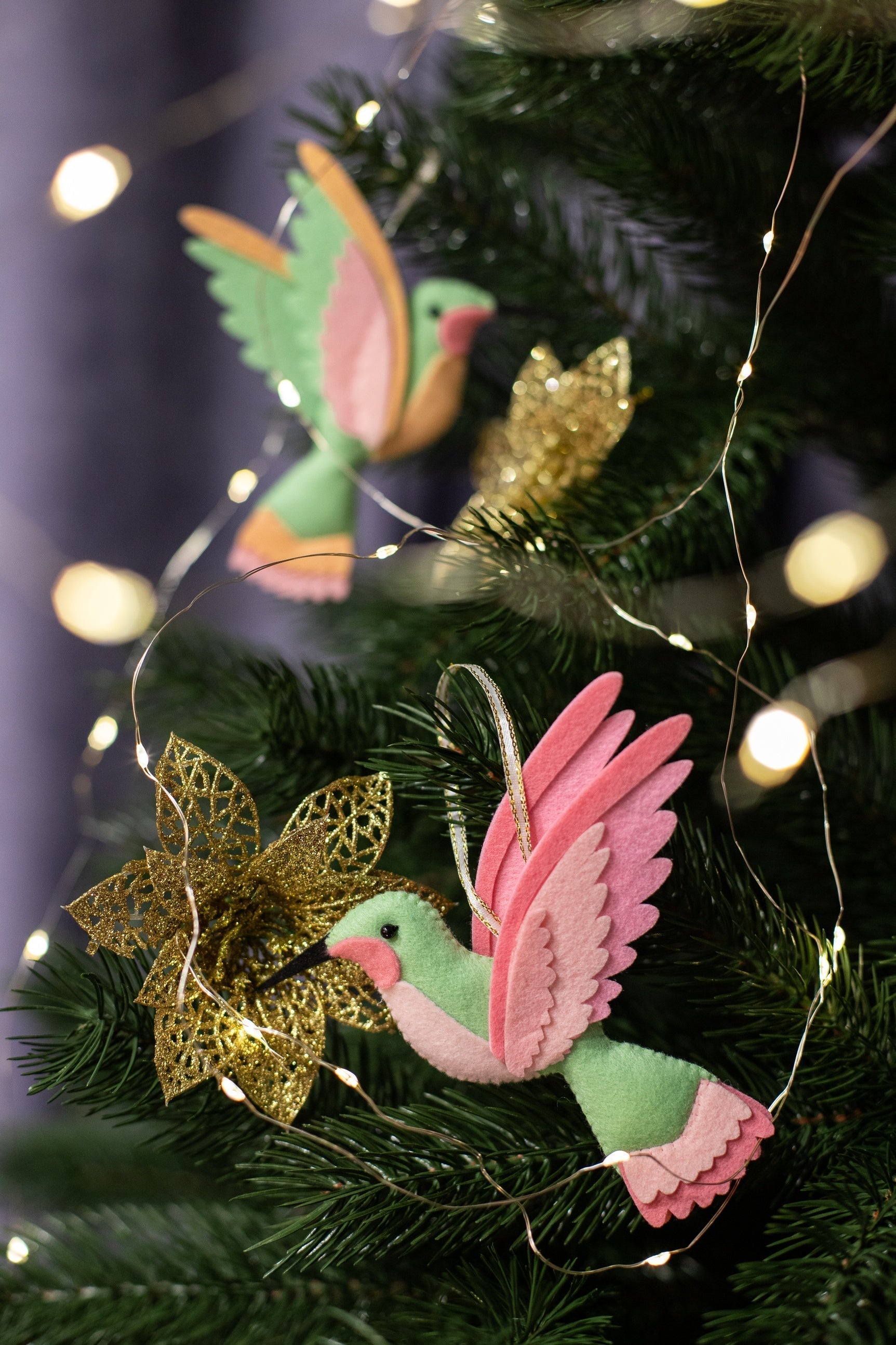 Handmade felt hummingbird ornament. The bird is mint green with a black beak. Its wings are a blend of pink and light pink. The background is a blurred Christmas tree with glowing festive lights.