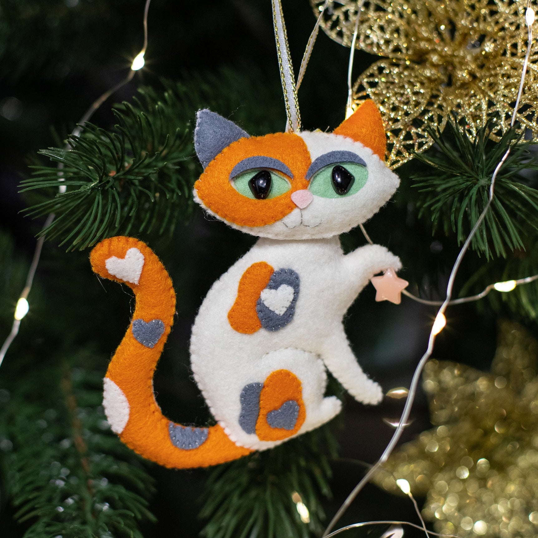 A white felt cat ornament with orange and grey patches, a small yellow bow, and a curled tail 