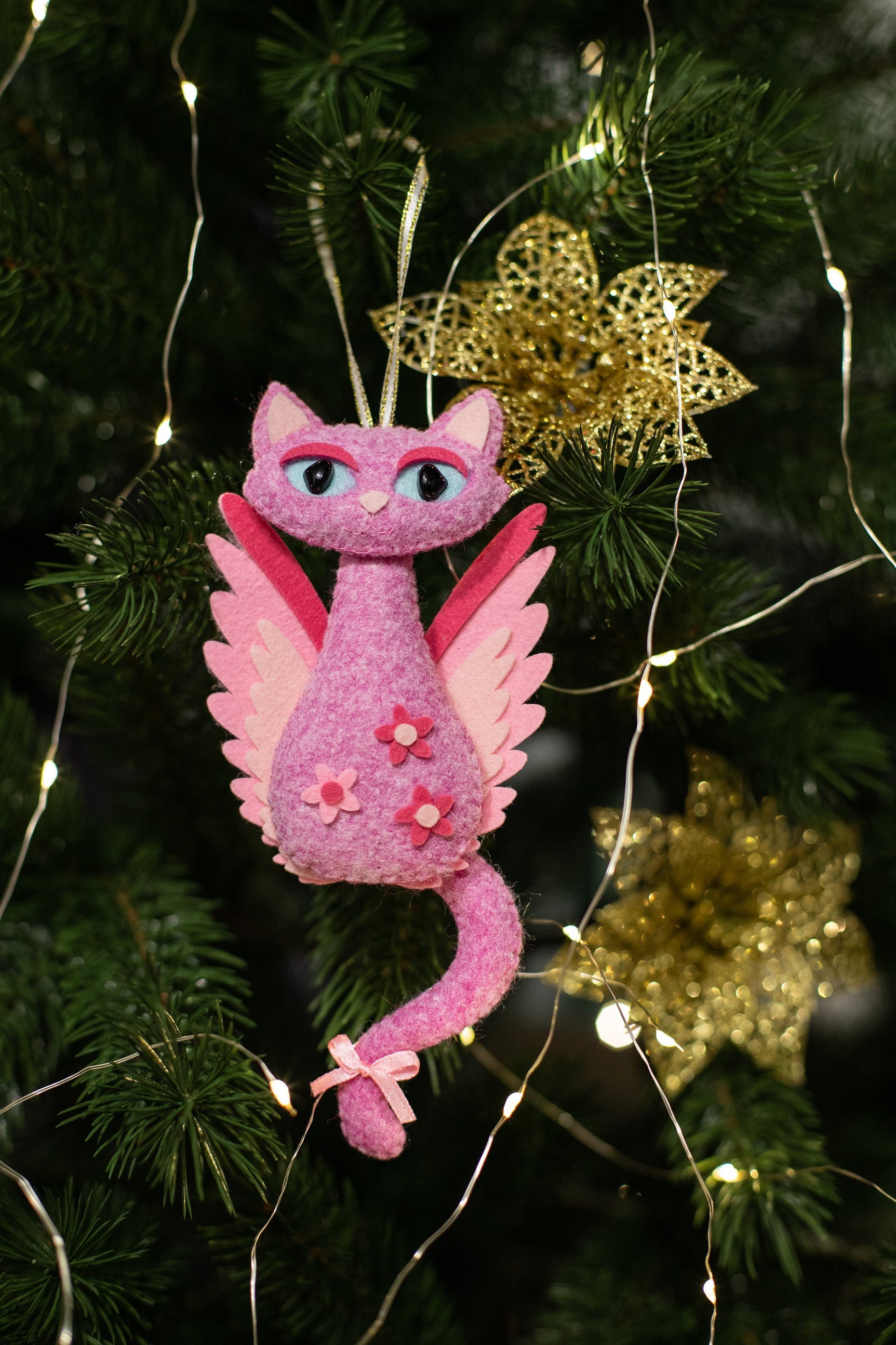 Pink felt cat Christmas ornament with wings and a gold halo. The cat has pink and white wings and a long tail. The cat has pink flowers on its body.