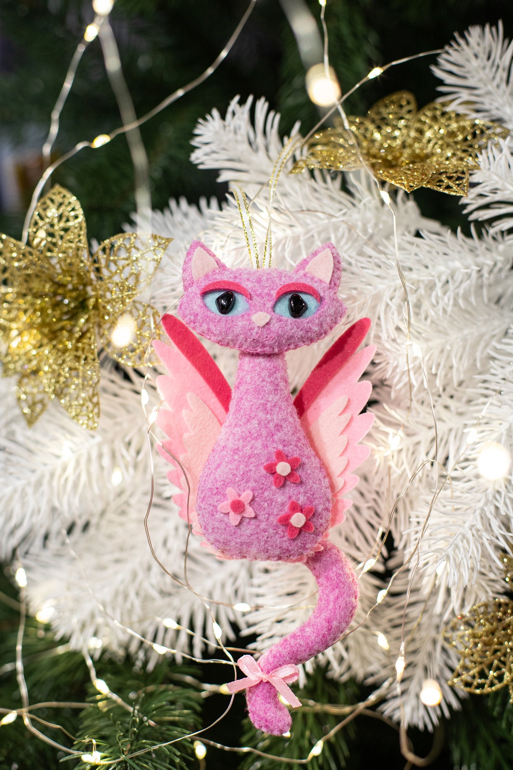 Pink felt cat Christmas ornament with wings and a gold halo. The cat has pink and white wings and a long tail. The cat has pink flowers on its body.