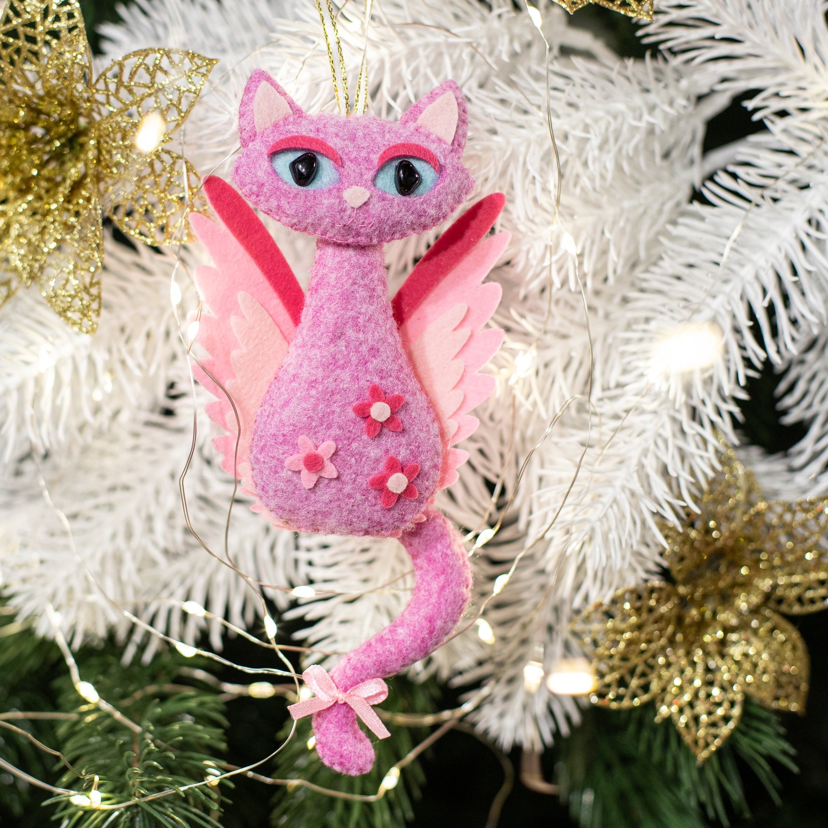 Pink felt cat Christmas ornament with wings and a gold halo. The cat has pink and white wings and a long tail. The cat has pink flowers on its body.