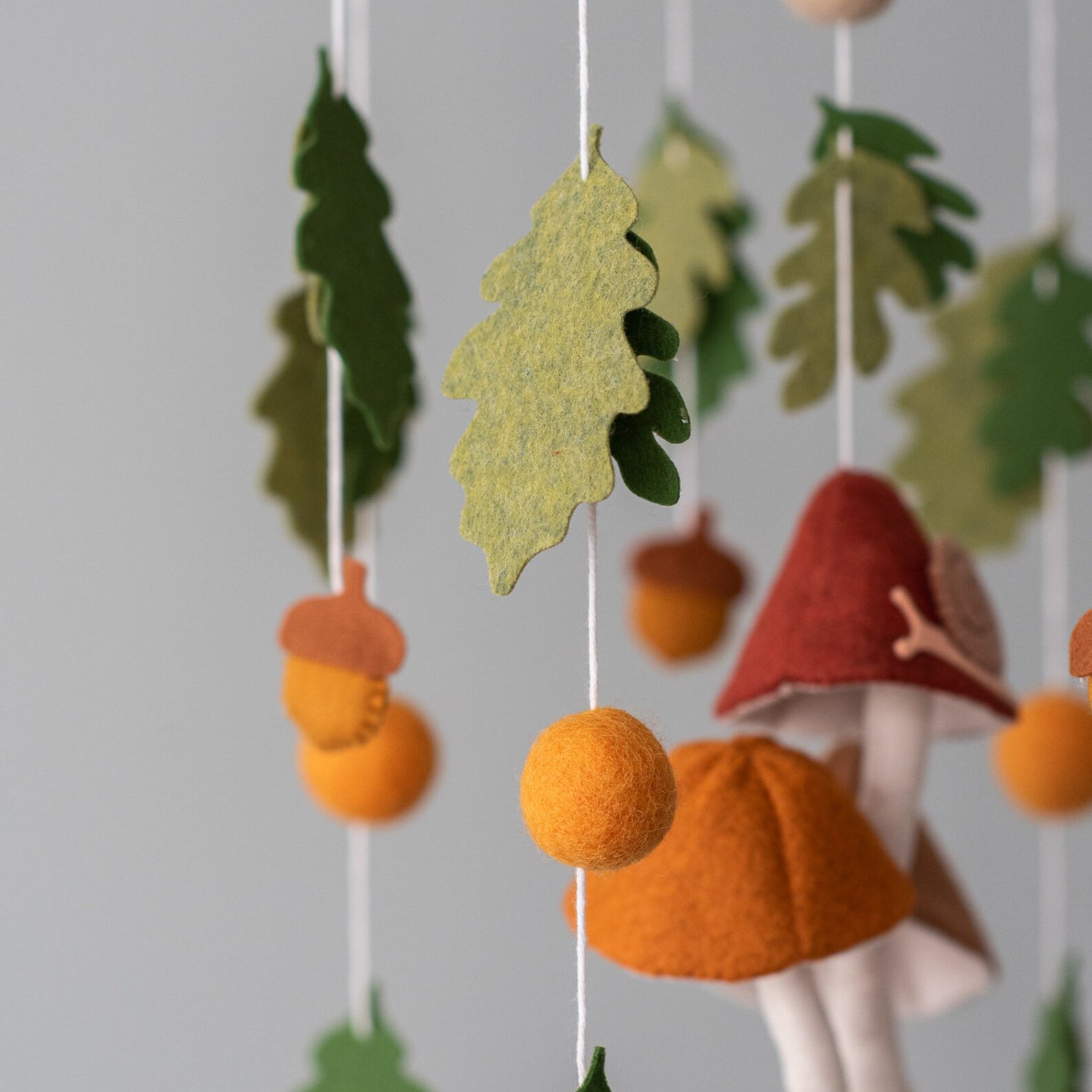 A handcrafted baby mobile featuring three large felt mushrooms in red, orange, and brown hanging in the center, with a small snail on one. Surrounding them are green oak leaves, orange acorns, and felt balls suspended from a wooden hoop.